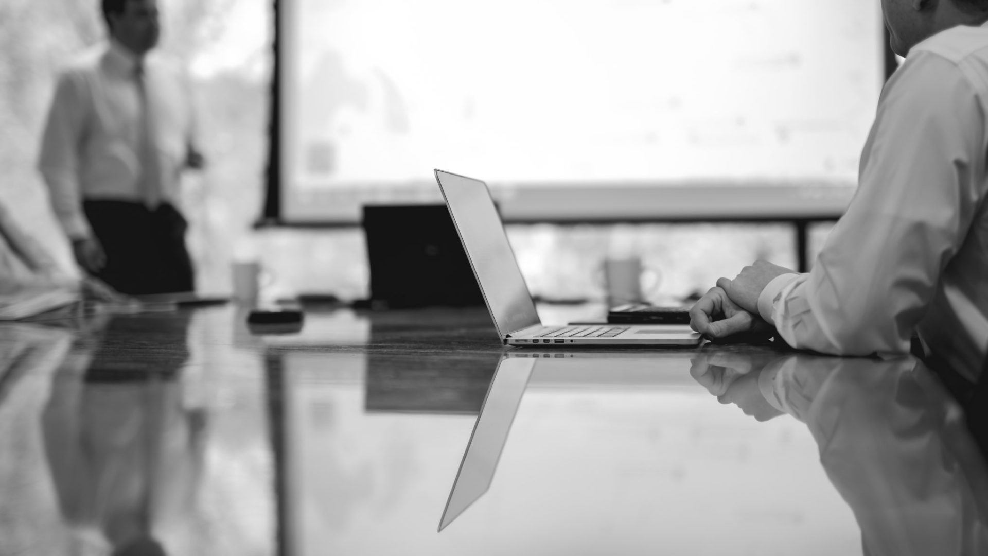 black and white photo of person sitting at table with laptop looking at projector